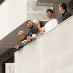 Cinco personas se asoman a un moderno balcón blanco con barandilla de rejilla en un bloque de viviendas públicas del Ayuntamiento de Rivas. Dos saludan y sonríen, mientras los otros miran al frente. El fondo muestra un edificio de ladrillo y otros balcones.