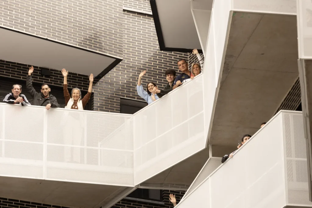 Un grupo de personas, de pie en distintos niveles de una escalera exterior adosada a un edificio de ladrillo, saludan a la cámara: residentes que celebran las nuevas viviendas públicas promovidas por el Ayuntamiento de Rivas.