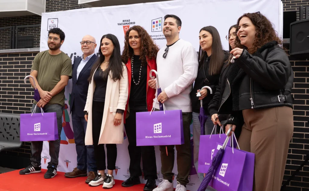 Un grupo de siete personas se para en una alfombra roja frente a un telón de fondo de marca en Rivas, sosteniendo bolsas de regalo moradas y sonriendo a la cámara durante un acto de promoción de viviendas públicas y alquiler para jóvenes.