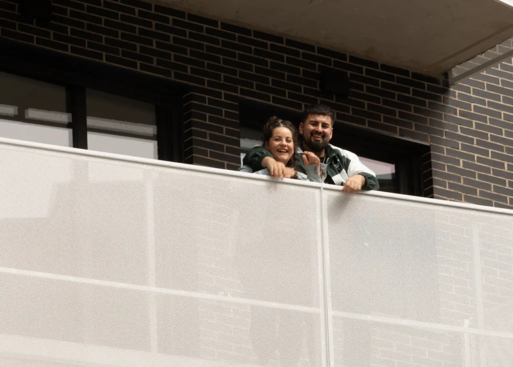Dos personas están de pie, una al lado de la otra, asomadas a la barandilla de un balcón, sonriendo. El fondo muestra una pared de ladrillo y ventanas de viviendas públicas del Ayuntamiento de Rivas, destinadas a alquiler para jóvenes.