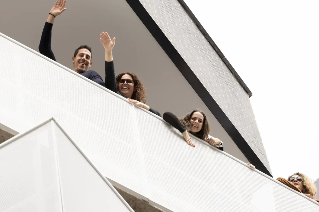 Tres personas se asoman a un balcón de unas modernas viviendas públicas, sonriendo y saludando, mientras una cuarta persona es parcialmente visible en el borde. El edificio presenta barandillas blancas y líneas geométricas, parte de una iniciativa del Ayuntamiento de Rivas de alquiler para jóvenes.