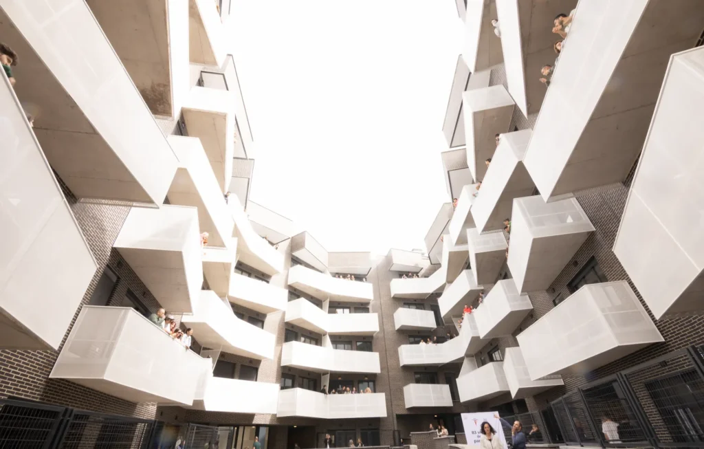Vista desde arriba de un moderno edificio de apartamentos con balcones angulares blancos, parte de las viviendas públicas del Ayuntamiento de Rivas, en cuyos balcones se ve a varios jóvenes disfrutando de un alquiler para jóvenes. El cielo es luminoso y está sobreexpuesto.