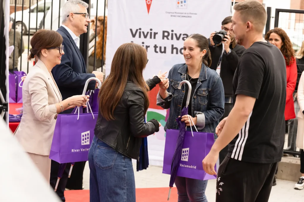 Varias personas intercambian bolsas de regalo moradas y se dan la mano. Al fondo se ve una pancarta del Ayuntamiento de Rivas, lo que sugiere que se trata de un acto relacionado con viviendas públicas o alquileres para jóvenes.