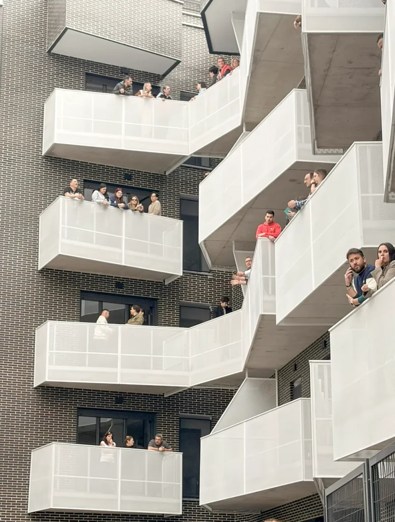 Varias personas de pie o apoyadas en las barandillas blancas de varios balcones de un moderno edificio de viviendas con paredes de ladrillo oscuro en Rivas, que presenta balcones blancos escalonados, un ejemplo de viviendas públicas y alquiler para jóvenes.
