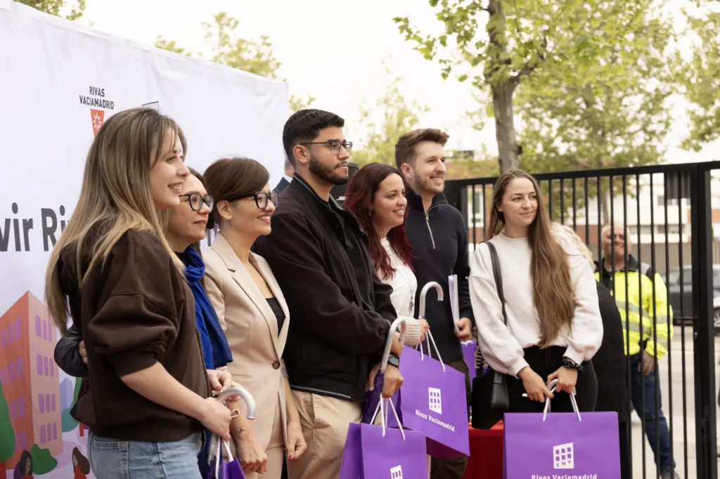 Un grupo de siete personas posa al aire libre delante de una pancarta de marca y una valla negra, con bolsas de regalo moradas y posando para una foto del acto de entrega de las 83 viviendas.