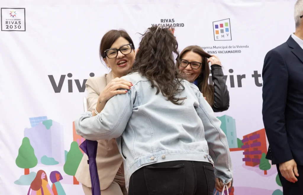 Dos mujeres con gafas se saludan con un abrazo en un acto al aire libre apoyado por el Ayuntamiento de Rivas. Otra persona permanece de pie a la derecha, parcialmente fuera de cuadro. Al fondo se ve una pancarta con logotipos e ilustraciones.