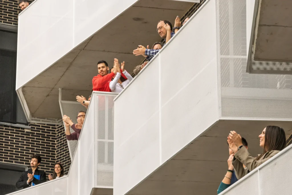 La gente aplaude de pie en los balcones de un edificio de varios pisos con barandillas de malla blanca, posiblemente celebrando las nuevas viviendas públicas del Ayuntamiento de Rivas. Los residentes parecen sonreír y aplaudir mientras miran hacia el exterior.