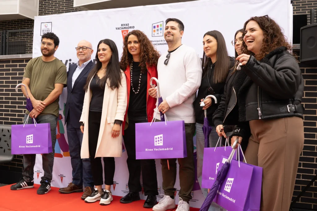 Un grupo de ocho personas permanecen juntas en una alfombra roja, sonriendo y sosteniendo bolsas de regalo moradas frente a un telón de fondo con la marca del Ayuntamiento de Rivas Vaciamadrid, que ha hecho entrega de 83 viviendas para jóvenes.