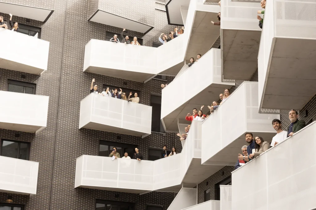 Varias personas miran desde varios balcones blancos de un moderno edificio de apartamentos de ladrillo oscuro en Rivas. La mayoría de los balcones tienen grupos de personas, que conforman los beneficiarios del acto de entrega de las 83 viviendas celebrado el pasado sábado.