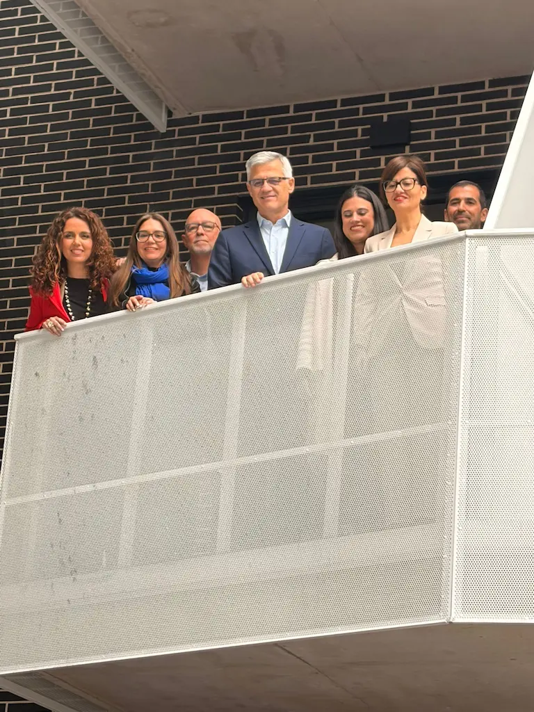 Siete personas, cuatro mujeres y tres hombres, permanecen juntas en un moderno balcón con barandilla de malla metálica en Rivas, posando para una foto de grupo frente a un oscuro muro de ladrillo que promociona viviendas públicas y alquiler para jóvenes.