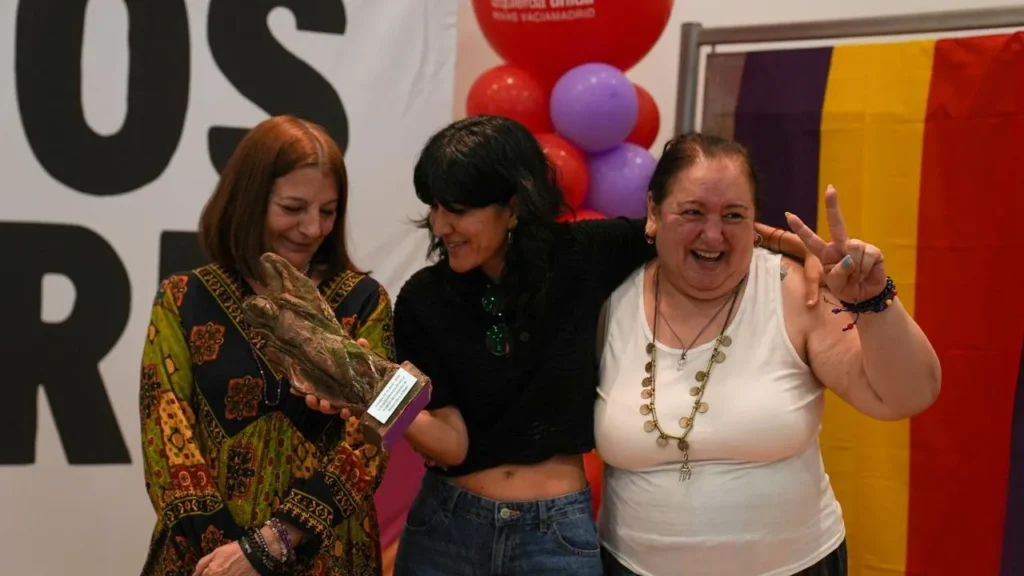 Tres mujeres sonríen juntas en las Jornadas Republicanas de Rivas; una sostiene una escultura de madera, otra hace el signo de la paz. Detrás de ellas hay globos multicolores, una gran pancarta y una bandera con los colores del arco iris entre 500 personas.