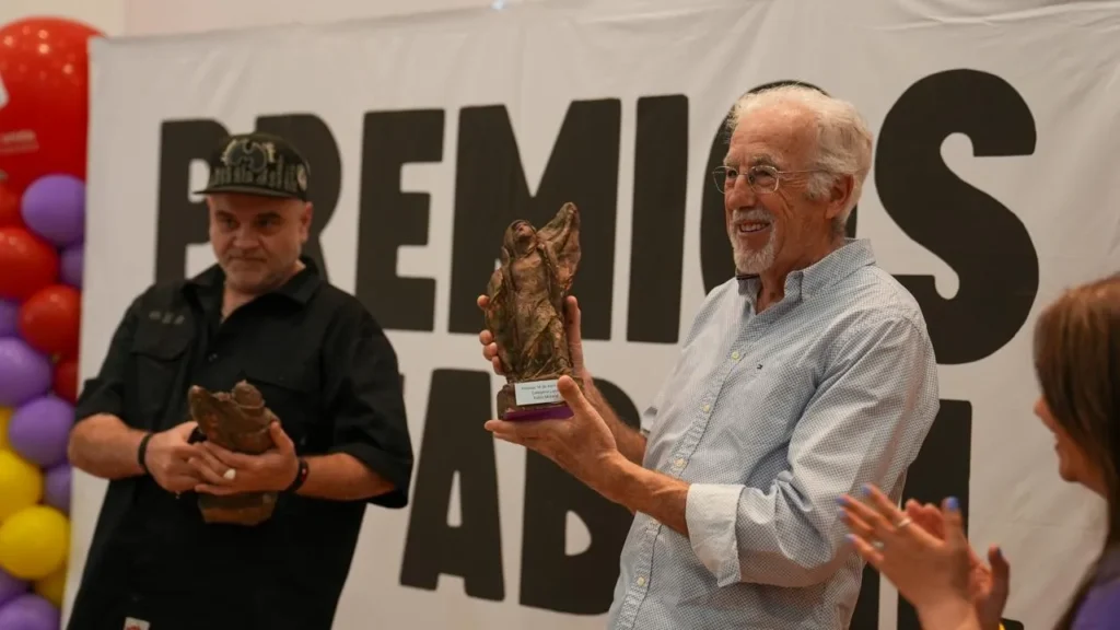 Dos hombres de pie delante de un cartel que dice PREMIOS AB, sosteniendo estatuas, mientras una mujer a la derecha aplaude. Globos de colores llenan el fondo, celebrando con 500 personas durante el evento Jornadas Republicanas en Rivas.