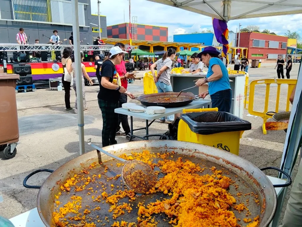 Una gran cacerola con restos de arroz en primer plano. La gente sirve comida bajo un toldo, con edificios de colores y un animado escenario con oradores y artistas al fondo.