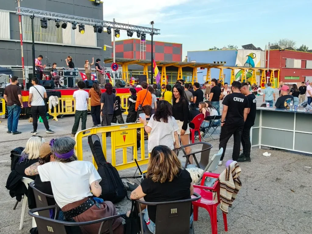 Unas 500 personas se reúnen en Rivas para disfrutar de un concierto al aire libre. Algunos se sientan en sillas detrás de una barricada amarilla, mientras que otros permanecen de pie o caminan, mientras el equipo y el personal del evento dirigen la escena con edificios al fondo.