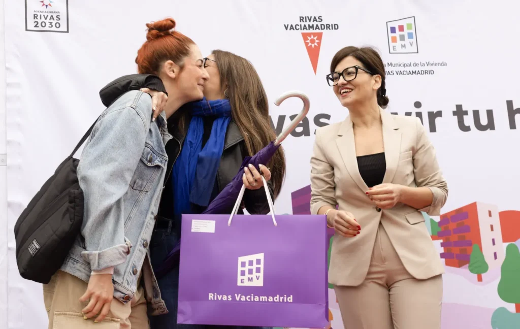 Tres mujeres ante una pancarta del Ayuntamiento de Rivas Vaciamadrid. Dos se saludan con un beso en la mejilla, mientras la tercera, con una carpeta morada y un paraguas en la mano, sonríe a su lado.