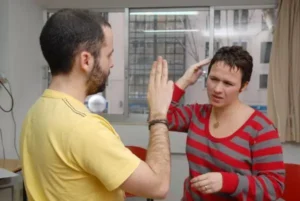 Dos personas se encuentran frente a frente en un interior. La persona de la izquierda levanta una mano, mientras que la de la derecha, con camisa roja a rayas, hace un gesto cerca de la cabeza, posiblemente utilizando Lengua de Signos Española. Al fondo se ven una ventana y sillas.