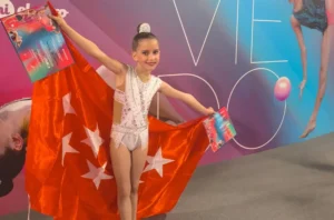 Adhara Rivas, una joven con un maillot blanco, sostiene una gran bandera roja con estrellas blancas y dos certificados. Se alza orgullosa ante un colorido telón de fondo con grandes letras, imágenes de gimnastas y símbolos de gimnasia rítmica.