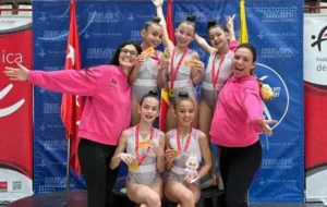 Seis jóvenes gimnastas alevines con leotardos y dos mujeres con sudaderas rosas posan juntas, sonrientes y sosteniendo medallas, celebrando como campeonas de Madrid en una competición de gimnasia en Rivas.