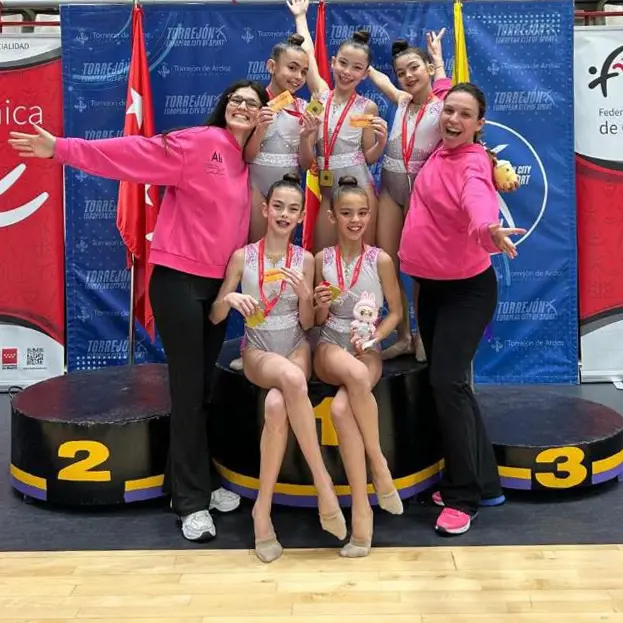 Seis gimnastas alevines de Rivas con maillots a juego y dos mujeres con chaquetas rosas posan en un podio de ganadores con medallas y peluches, sonriendo a la cámara. El telón de fondo muestra carteles y banderas de campeonas de Madrid.