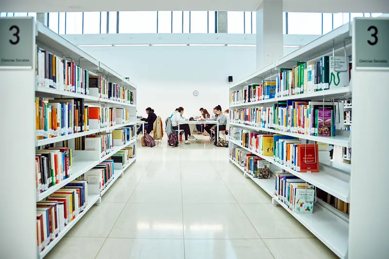 Hileras de estanterías en una iluminada biblioteca de Rivas, con varias personas sentadas en mesas al fondo, leyendo o estudiando durante época de exámenes y consultando horarios de bibliotecas.