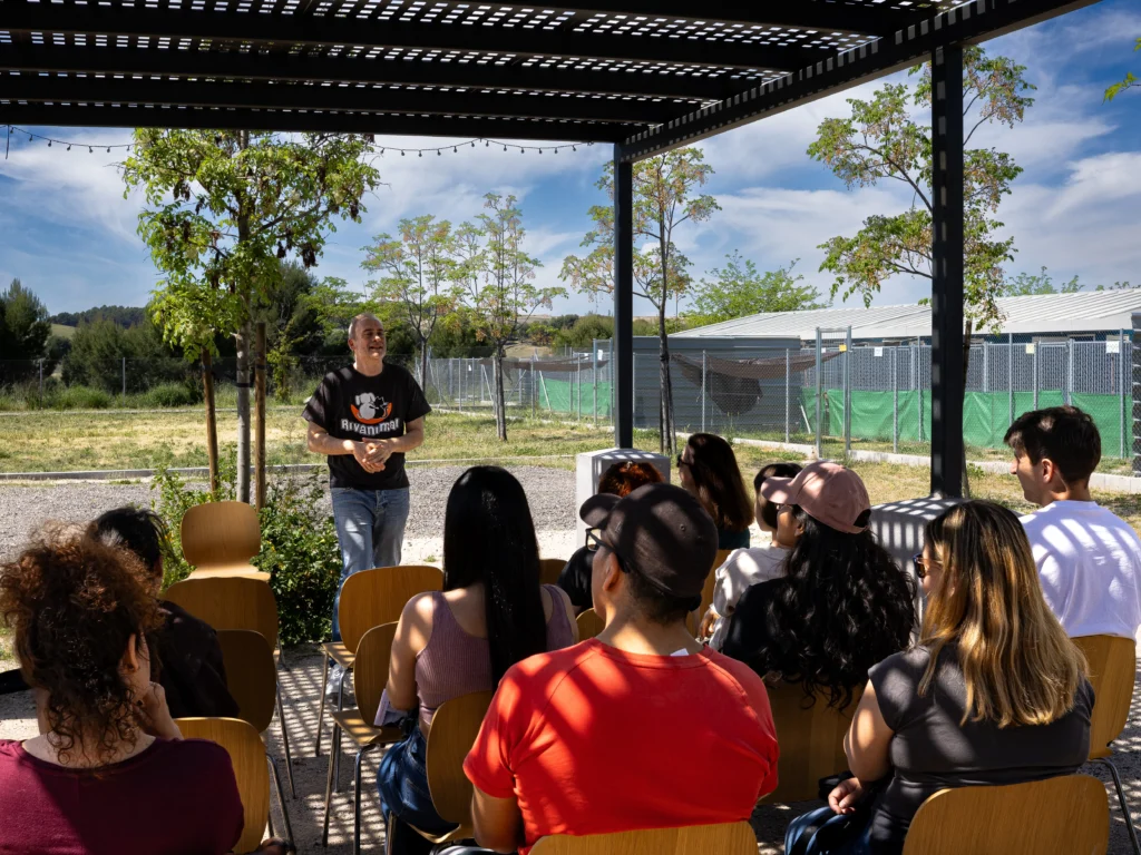 Una persona habla de pie ante un público sentado al aire libre bajo una pérgola en un acto de Rivanimal, con árboles, zona de césped y edificios vallados de fondo. El público escucha atentamente mientras se tratan temas como las adopciones y el voluntariado.