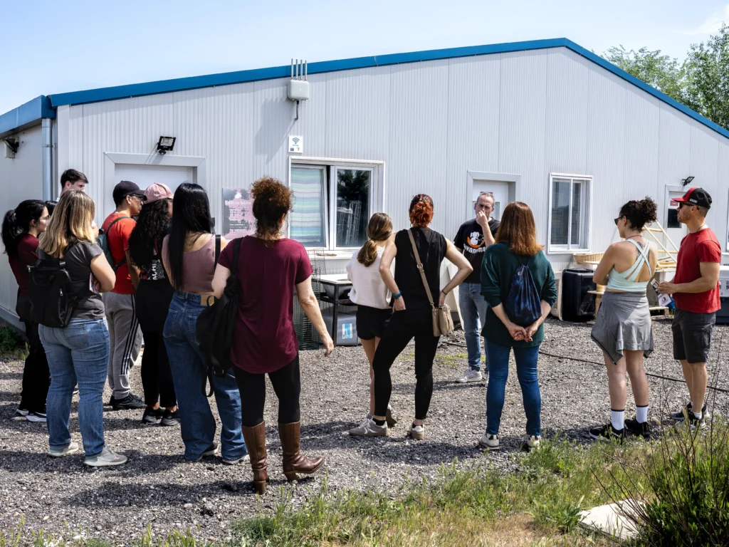 Un grupo de personas permanece de pie frente a un edificio modular blanco con tejado azul, escuchando a un orador de Rivanimal. El grupo, vestido de manera informal, parece enfrascado en una discusión sobre voluntariado y adopciones en este día soleado.