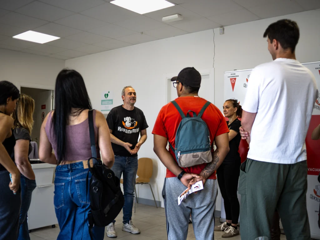 Un grupo de personas, algunas con mochilas, permanecen de pie en un semicírculo en el interior, escuchando a un hombre con camiseta negra que habla sobre el voluntariado en la entidad protectora. El escenario parece una oficina o una sala de espera.
