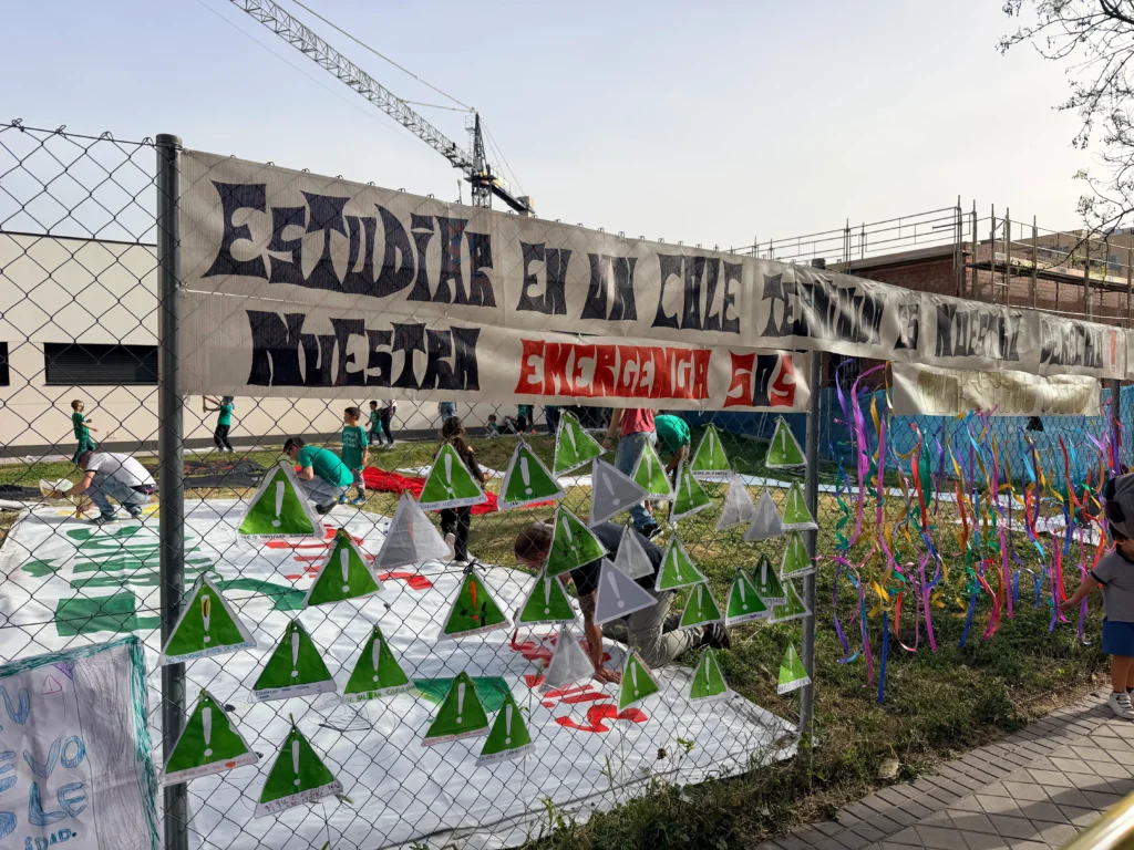 Una valla de alambre en el CEIP Mercedes Vera muestra una pancarta en español y adornos de papel. Detrás de la valla, personas con camisas verdes trabajan cerca de una gran lámina pintada en el suelo.