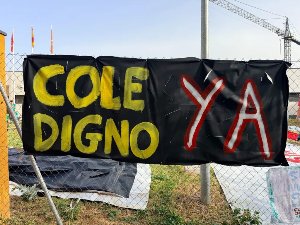 Una pancarta negra fijada a una valla del CEIP Mercedes Vera muestra las palabras COLE DIGNO en amarillo y YA en grandes letras rojas y blancas, con materiales de construcción en el fondo.