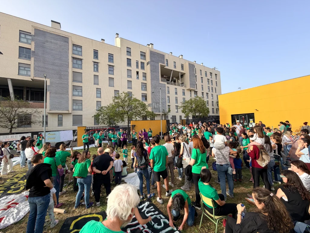 Un nutrido grupo de personas, muchas de ellas con camisetas verdes, se reúnen al aire libre en una zona de césped frente a un moderno edificio de apartamentos y un muro amarillo durante un encierro diurno para protestar por el retraso de las obras en el CEIP Mercedes Vera.