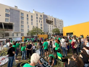 Un nutrido grupo de personas, muchas de ellas con camisetas verdes, se reúnen al aire libre en una zona de césped frente a un moderno edificio de apartamentos y un muro amarillo durante un encierro diurno para protestar por el retraso de las obras en el CEIP Mercedes Vera.