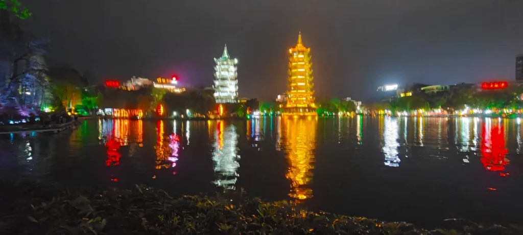 Dos pagodas iluminadas se alzan junto a un lago nocturno de Guilin, una blanca y otra dorada, con las luces de colores de los edificios circundantes y las torres reflejándose en la superficie del agua.