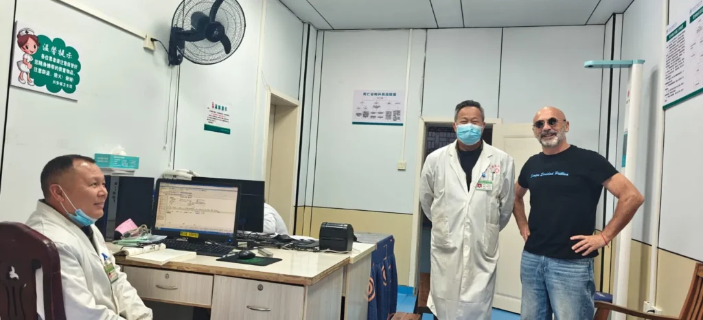 Tres hombres de pie en una consulta médica; dos con batas blancas de laboratorio y mascarillas, uno con camiseta negra y gafas de sol. Se ven historiales y equipos médicos, lo que sugiere que se trata de una clínica situada posiblemente en Shenzhen o en la cercana Macao.