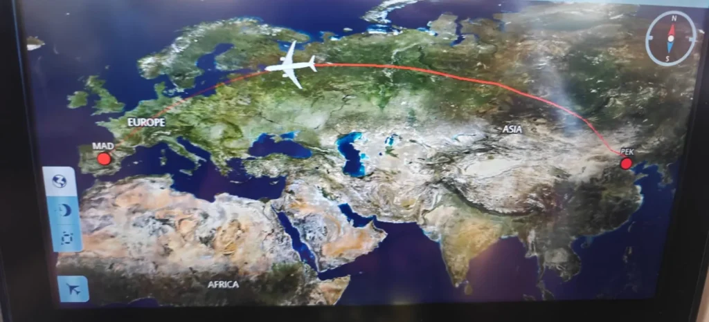 Una pantalla de mapa en vuelo muestra un avión que vuela de Madrid (MAD), España, a Pekín (PEK), China, con una línea de ruta curva roja a través de Europa y Asia. En la esquina superior derecha aparece una brújula y Hong Kong aparece cerca en el mapa.