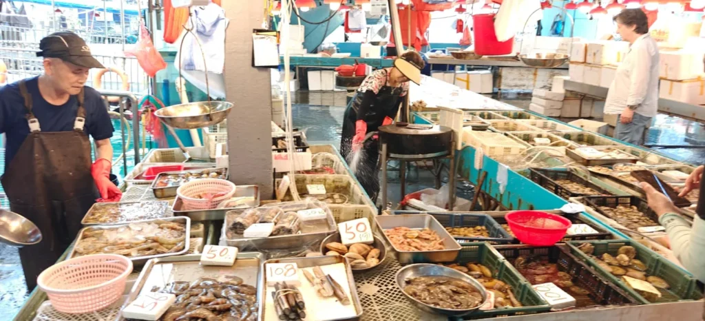 Un mercado de marisco con bandejas de diversos mariscos frescos expuestos, con las etiquetas de precio visibles, evoca recuerdos de Hong Kong. Hay tres trabajadores: uno a la izquierda vestido de azul, otro en el centro manipulando marisco y un tercero al fondo.
