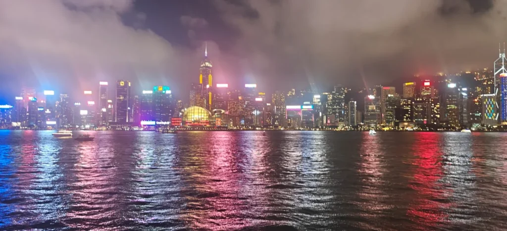 Un paisaje urbano nocturno de Hong Kong con rascacielos iluminados que reflejan luces de colores sobre el agua; las nubes flotan en el cielo sobre la ciudad, evocando recuerdos urbanos.