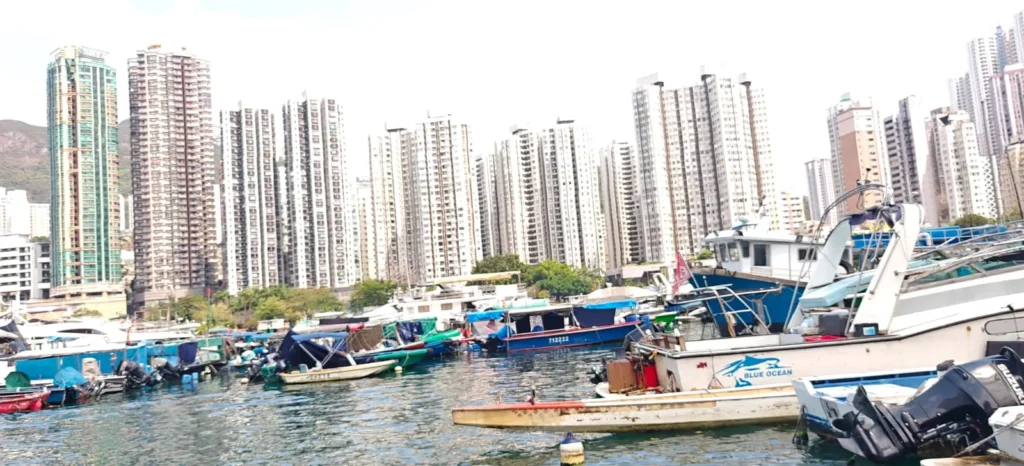 Varios barcos y buques pesqueros flotan en el agua en un ajetreado puerto de Hong Kong, con altos edificios de apartamentos residenciales muy juntos al fondo, bajo un cielo luminoso.