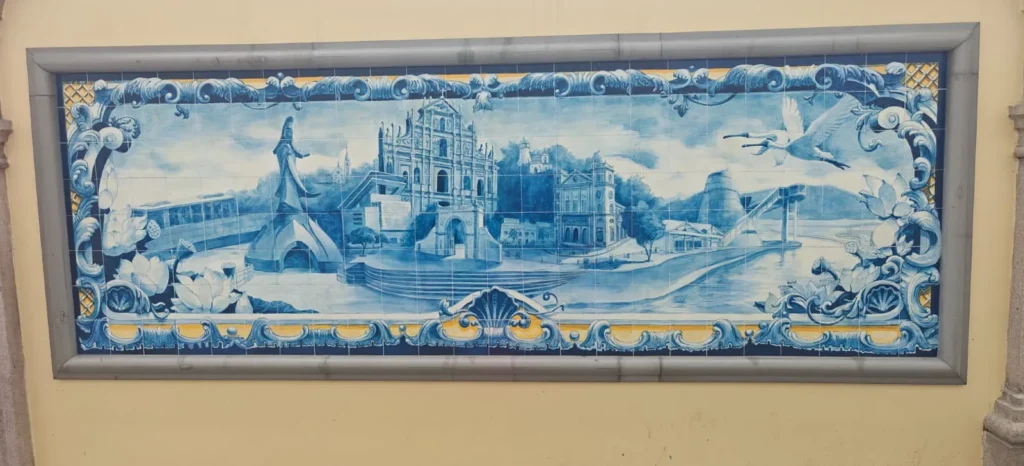 Un mural rectangular titulado "Memorias por China", realizado con azulejos azules y blancos, representa varios edificios, una iglesia, un puente, árboles, pájaros y motivos decorativos sobre una pared amarilla. Su borde presenta elementos azules y amarillos que recuerdan Hong Kong-Pekín-Rivas.