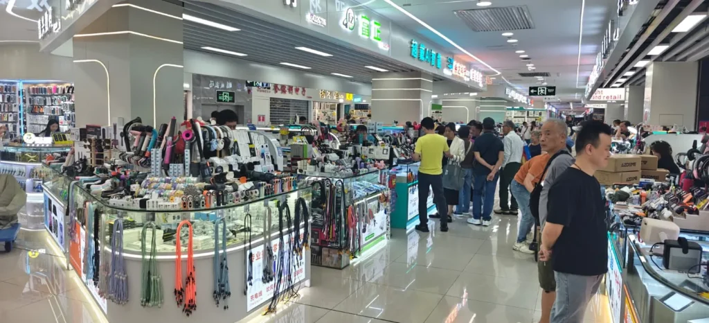 Un concurrido mercado de electrónica y accesorios de Hong Kong con mostradores de cristal, varios aparatos y accesorios de telefonía a la venta, y muchos compradores paseando o curioseando por pasillos bien iluminados.