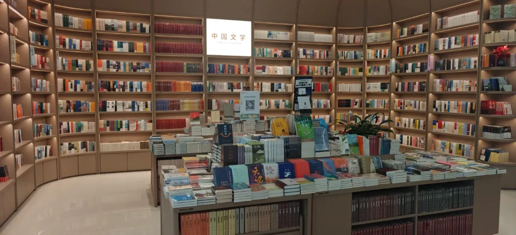 Una librería bien iluminada de Hong Kong con estanterías repletas de libros ordenados. Una mesa expositora central está repleta de libros, y un cartel con caracteres chinos se ilumina sobre las estanterías.