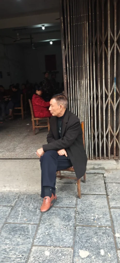 Un hombre con abrigo oscuro y zapatos marrones se sienta en una silla de madera en el exterior de un edificio con verja metálica en Pekín. Varias personas están sentadas dentro de la habitación poco iluminada del fondo.