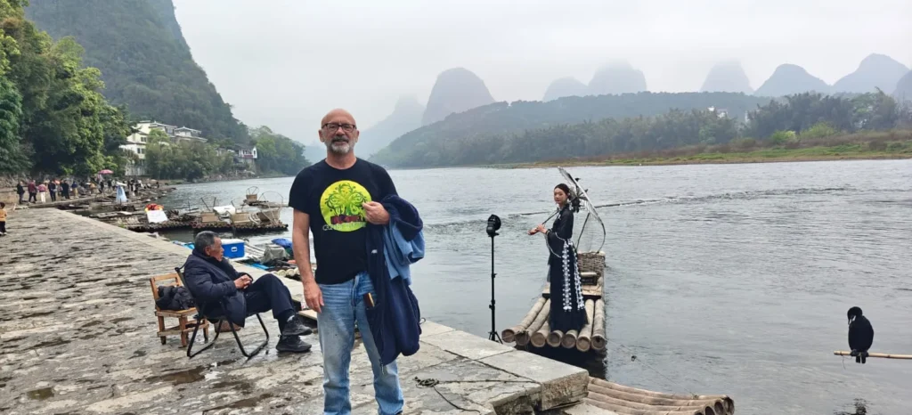 Un hombre en una pasarela de piedra cerca de un río en China, con montañas al fondo. Otras personas están sentadas o de pie junto al río, mientras que una mujer está de pie en una balsa de bambú donde un pájaro cormorán se posa en el borde.