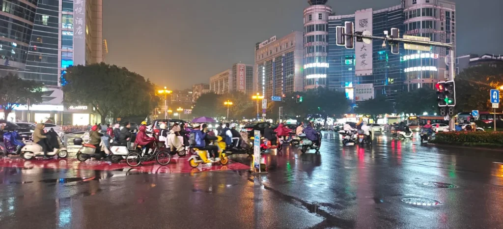 Un concurrido cruce urbano de Hong Kong por la noche con muchas personas en patinetes y bicicletas esperando en un semáforo. Altos edificios con letreros luminosos rodean las calles húmedas y reflectantes bajo un cielo nublado.