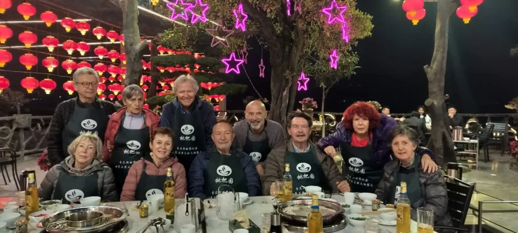 Un grupo de once adultos, algunos con delantales a juego, posan alrededor de una mesa redonda de comedor al aire libre cubierta de platos y bebidas. De los árboles cuelgan farolillos rojos y luces de estrellas rosas, que evocan recuerdos festivos de las noches de Hong Kong o Pekín.