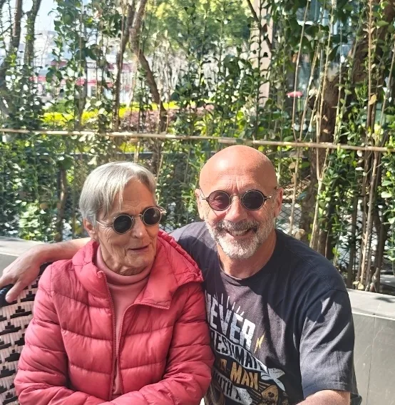 Una mujer mayor con una chaqueta rosa y un hombre mayor con una camiseta gráfica negra sentados uno al lado del otro en Pekín, ambos con gafas de sol redondas. El hombre rodea a la mujer con el brazo, con un follaje verde como telón de fondo.