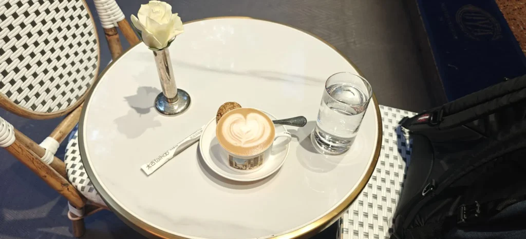 Una mesa redonda de mármol blanco sostiene un capuchino con arte latte, un biscotti, un vaso de agua, una rosa blanca en un jarrón, una servilleta y una bolsa negra en una silla estampada a su lado: recuerdos de un café de Hong Kong.