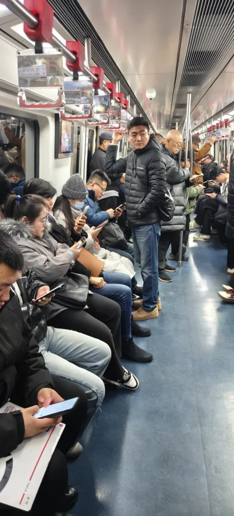 Pasajeros sentados y de pie en un vagón de metro abarrotado, muchos con chaquetas de invierno y absortos en sus teléfonos. El vagón, brillantemente iluminado, recuerda los desplazamientos al trabajo en Pekín, con suelos azules y pasamanos que se extienden por encima de los asientos: recuerdos urbanos en movimiento.