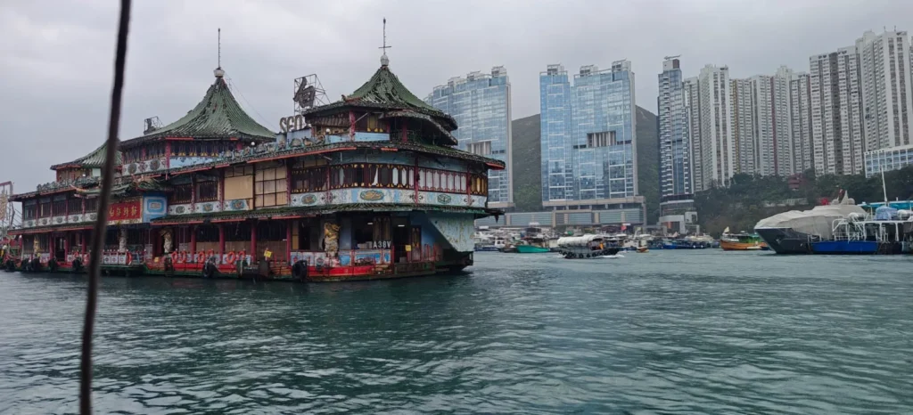 Un restaurante flotante de ornamentada arquitectura tradicional china se asienta sobre el agua en Shenzhen, con modernos rascacielos y otros barcos de fondo bajo un cielo nublado.