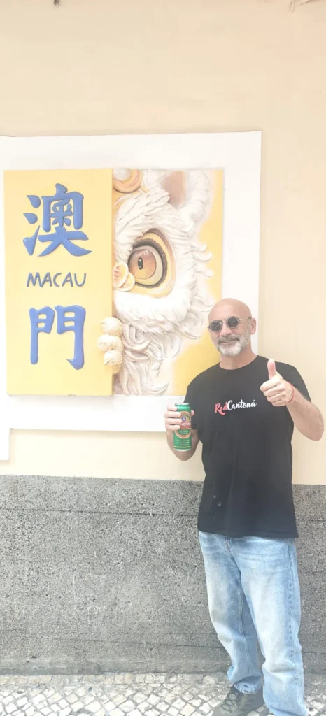 Un hombre con gafas de sol y camiseta negra, de pie junto a un mural con caracteres chinos y la palabra "Macao", sostiene una lata y levanta el pulgar. El mural, encontrado cerca de Shenzhen, representa parte de la cara de un león blanco.