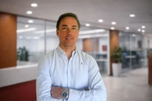 Un hombre de pelo castaño corto con una camisa azul claro abotonada está de pie con los brazos cruzados en una moderna oficina de España con paredes de cristal, plantas en macetas y luces en el techo, que reflejan el espíritu innovador de Fazil Crypto y la normativa MiCA.